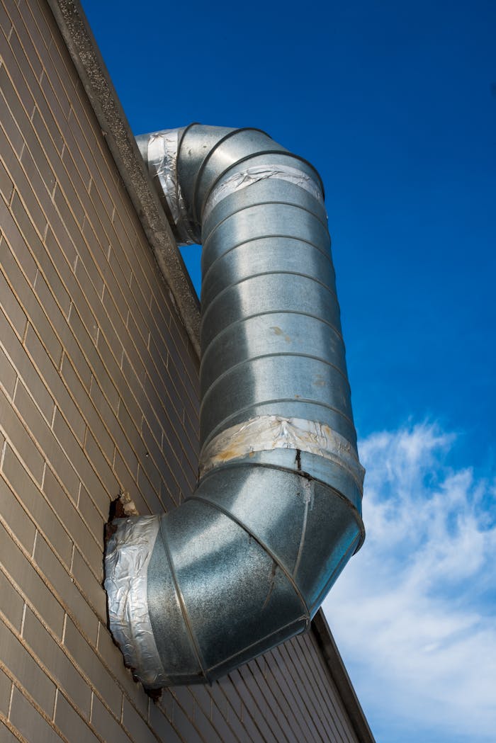 about-01 A large industrial air duct attached to a brick wall under a bright blue sky.